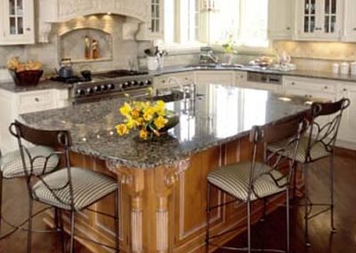 A stylish kitchen features a large granite island with intricate wooden supports. Four cushioned bar stools are placed around the island. The space has white cabinetry, stainless steel appliances, and a decorative range hood. A vase with yellow flowers is on the island.