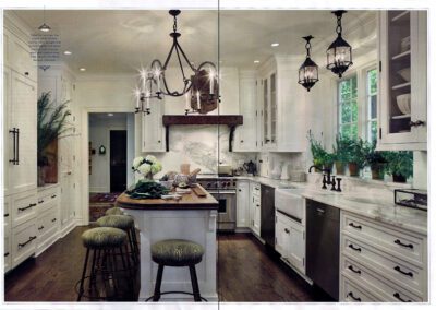 A spacious, rustic-themed kitchen with white cabinetry, marble countertops, and stainless steel appliances. A wooden island with two cushioned stools is centered. The room features pendant lighting, large windows, and various plants for decor.