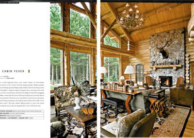 A magazine spread features a cozy log cabin interior with large windows showcasing a forest view. The warm living space includes wooden furniture, a stone fireplace, a mounted deer head, and a chandelier. The left page offers a text description and article title, "Cabin Fever.