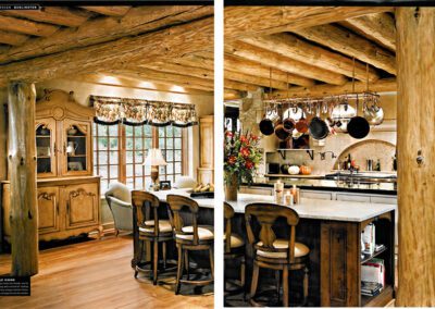 A rustic kitchen and dining area with wooden beams and pillars. The kitchen has a large island with stools, an oven, and hanging pots. The dining area features a wooden hutch, cushioned chairs, and a window with floral curtains, creating a cozy cabin atmosphere.