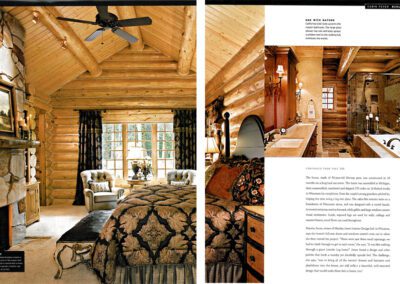 A luxurious log cabin interior featuring a cozy bedroom with a large bed, patterned bedding, and plush seating area in front of a stone fireplace. The room also includes a bathtub with ornate fixtures, wooden beams, a ceiling fan, and large windows with picturesque views.