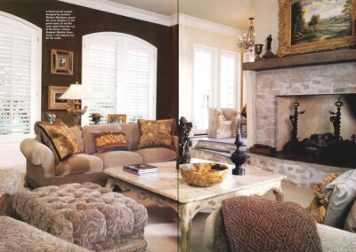 A cozy living room with beige and brown tones, featuring plush sofas adorned with patterned cushions, a tufted ottoman, a stone fireplace with a framed painting above it, a decorative bowl on a wooden coffee table, and large windows with white shutters.