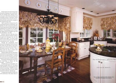 A cozy, old-world style kitchen with floral curtains, white cabinetry, and dark wooden floors. A decorative chandelier hangs above a wooden dining table set with elegant dinnerware and floral centerpiece. The room is filled with warm, inviting light.