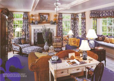 A cozy living room with floral-themed decor. It features a wicker sofa, a white wooden desk, a fireplace with a white mantle, multiple patterned sofas and chairs, and large windows with floral curtains. Lamps, vases, and framed artwork add to the room's charm.