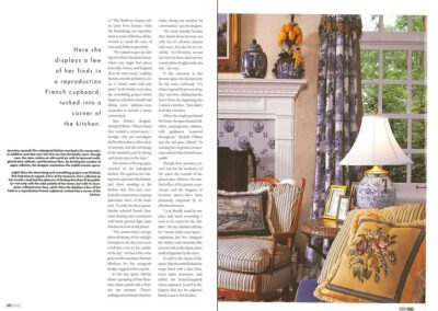 A magazine spread features an elegant living room with a cozy seating area adorned with blue and yellow floral patterns, antique porcelain vases, framed wall art, and detailed curtains. A table lamp is placed on a side table, illuminating the space. Text is visible on the left page.