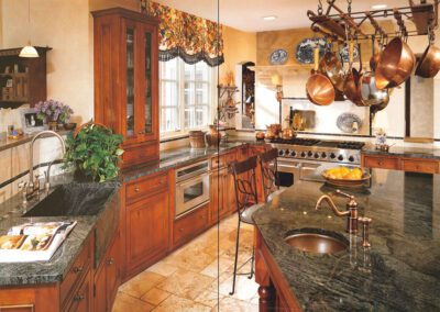 A spacious, rustic kitchen featuring wooden cabinets, granite countertops, and stainless steel appliances. Copper pans hang above a large island with a sink. A pot of leafy green plant sits next to the main sink by the window, providing plenty of natural light.