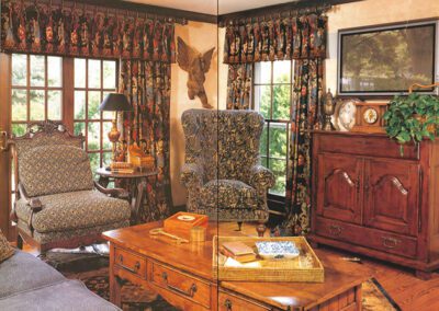 A cozy living room with patterned fabric chairs, a wooden coffee table, and a large TV mounted above a wooden cabinet. Decorative items include a clock, books, and a potted plant. Two large windows with heavy drapes bring in natural light, revealing greenery outside.